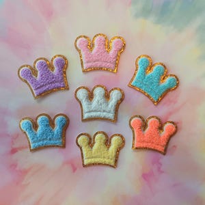 May include: Seven colorful crown patches in shades of purple, pink, blue, white, yellow, and orange. Each crown has a textured appearance and is outlined with gold glitter. The patches are arranged on a soft, multi-colored background.