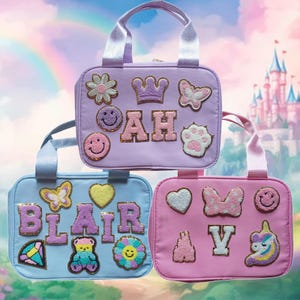 May include: Three pastel-colored tote bags with various embroidered patches. The bags are light blue, lavender, and pink. The patches include a crown, a paw print, a butterfly, a heart, a unicorn, a flower, and a smiley face. The bags are personalized with the names "Blair", "AH", and "V".