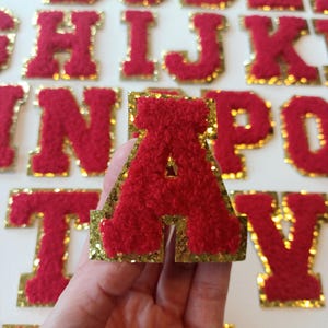 May include: Red felt letter patches with gold glitter trim. The letter A is being held in a hand. The letters are in a variety of sizes.