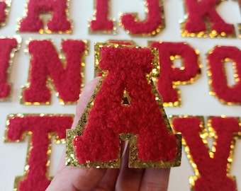 May include: Red felt letter patches with gold glitter trim. The letter A is being held in a hand. The letters are in a variety of sizes.
