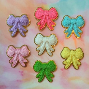 May include: Seven colorful felt bows with gold glitter trim. The bows are in shades of pink, purple, blue, white, yellow, and green.