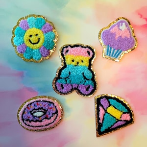 May include: A collection of colorful, textured patches in various shapes. Includes a flower with a smiling face, a teddy bear, a cupcake, a donut, and a geometric design, all with gold glitter borders.