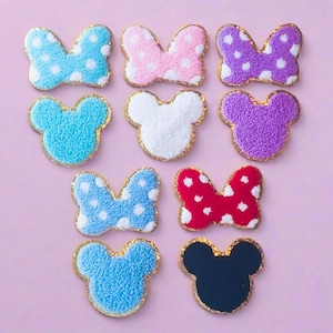 May include: An assortment of colorful, plush Minnie Mouse-themed patches and appliques. The collection includes bows with white polka dots in blue, pink, and purple, and Mickey Mouse head shapes in blue, white, purple, and black, all with gold trim.