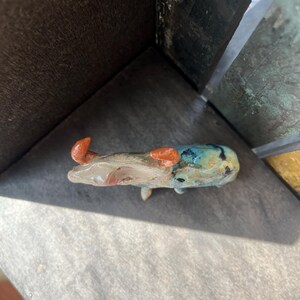 K&ouml;nnte beinhalten: Eine kleine Keramikfigur eines Stiers mit gl&auml;nzender Oberfl&auml;che. Der K&ouml;rper weist eine Mischung aus gesprenkelten Blau-, Gr&uuml;n- und Braunt&ouml;nen auf, w&auml;hrend die beiden geschwungenen H&ouml;rner eine markante, texturierte rot-orange Farbe haben.