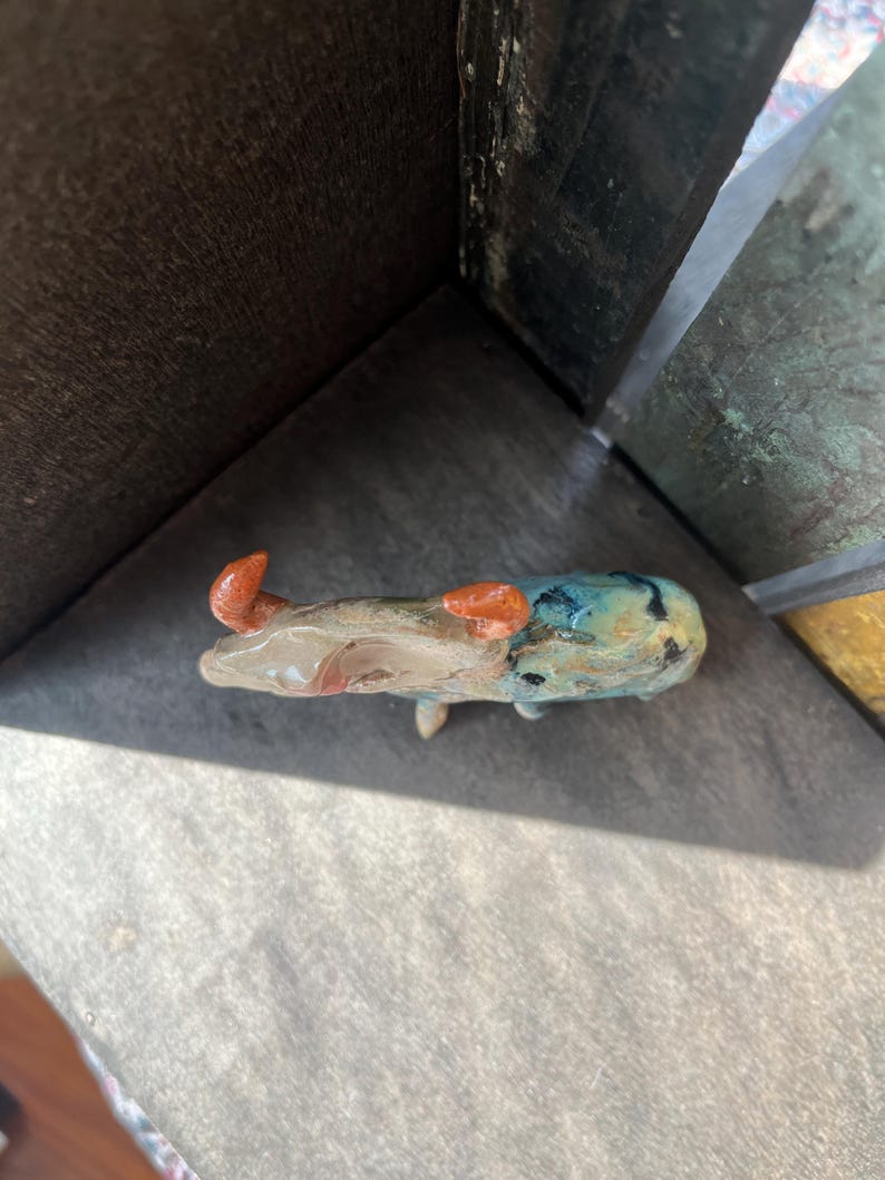 K&ouml;nnte beinhalten: Eine kleine Keramikfigur eines Stiers mit gl&auml;nzender Oberfl&auml;che. Der K&ouml;rper weist eine Mischung aus gesprenkelten Blau-, Gr&uuml;n- und Braunt&ouml;nen auf, w&auml;hrend die beiden geschwungenen H&ouml;rner eine markante, texturierte rot-orange Farbe haben.