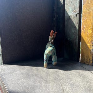 K&ouml;nnte beinhalten: Eine kleine Tierfigur aus Keramik mit einer gesprenkelten t&uuml;rkisfarbenen und blauen Glasur sowie einem braunen Kopf und Ohren. Das vierbeinige Wesen ist von hinten zu sehen und zeigt seine glatte, gl&auml;nzende Oberfl&auml;che und kompakte Form.