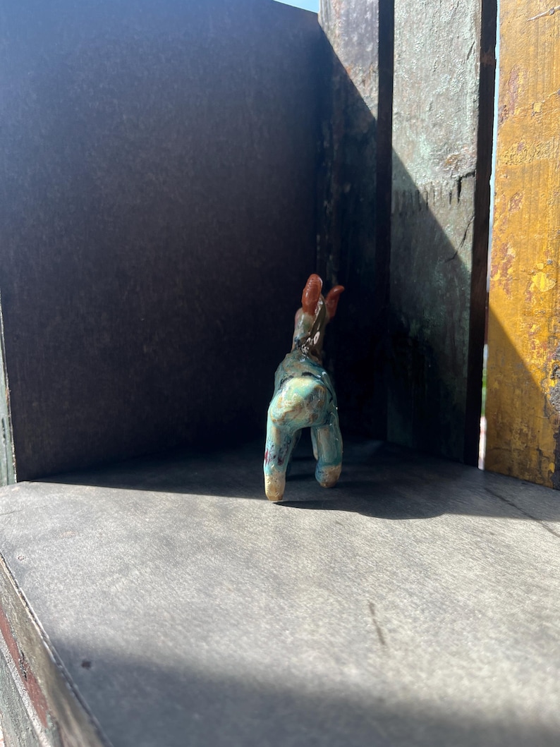 K&ouml;nnte beinhalten: Eine kleine Tierfigur aus Keramik mit einer gesprenkelten t&uuml;rkisfarbenen und blauen Glasur sowie einem braunen Kopf und Ohren. Das vierbeinige Wesen ist von hinten zu sehen und zeigt seine glatte, gl&auml;nzende Oberfl&auml;che und kompakte Form.