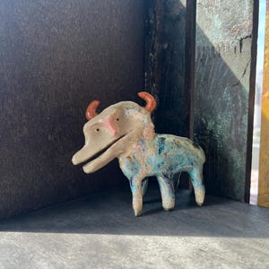 K&ouml;nnte beinhalten: Eine kleine, vierbeinige Tierfigur aus Keramik mit beigem Kopf, rosa Nase und zwei orangefarbenen H&ouml;rnern. Der K&ouml;rper ist mit einer gesprenkelten Glasur in Blau-, Gr&uuml;n- und Braunt&ouml;nen versehen. Das Wesen hat ein weit ge&ouml;ffnetes Maul und kleine, dunkle Augen.