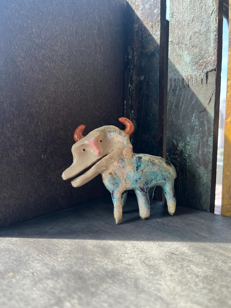 K&ouml;nnte beinhalten: Eine kleine, vierbeinige Tierfigur aus Keramik mit beigem Kopf, rosa Nase und zwei orangefarbenen H&ouml;rnern. Der K&ouml;rper ist mit einer gesprenkelten Glasur in Blau-, Gr&uuml;n- und Braunt&ouml;nen versehen. Das Wesen hat ein weit ge&ouml;ffnetes Maul und kleine, dunkle Augen.