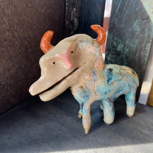 May include: A small ceramic animal figurine with a tan head and body featuring bright orange horns. The lower portion of the body is glazed in mottled shades of blue, white, and brown. The creature has a long snout, small eyes, and four legs.