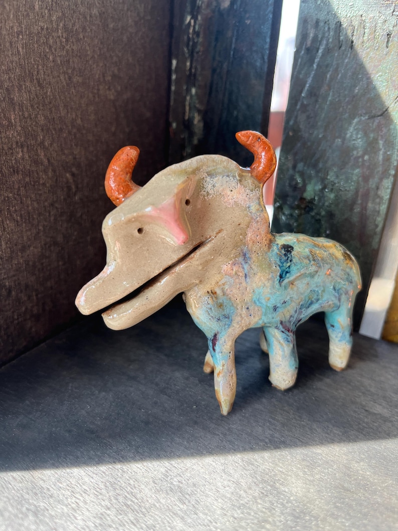 K&ouml;nnte beinhalten: Eine kleine Tierfigur aus Keramik mit beigem Kopf und K&ouml;rper sowie leuchtend orangefarbenen H&ouml;rnern. Der untere Teil des K&ouml;rpers ist in gesprenkelten Blau-, Wei&szlig;- und Braunt&ouml;nen glasiert. Das Wesen hat eine lange Schnauze, kleine Augen und vier Beine.