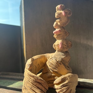 May include: A tan clay sculpture featuring a seated figure with large, textured hands and wings, supporting a vertical stack of three smaller, abstract faces with pink accents. The surface has visible etched lines and a rough, earthy texture.