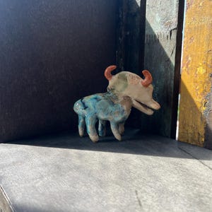 K&ouml;nnte beinhalten: Eine kleine Keramik-Tierfigur mit vier Beinen, einer langen Schnauze und zwei gebogenen orangefarbenen H&ouml;rnern. Der K&ouml;rper weist eine gesprenkelte Glasur in Blau-, Gr&uuml;n- und Cremet&ouml;nen auf, w&auml;hrend der Kopf haupts&auml;chlich cremefarben ist.