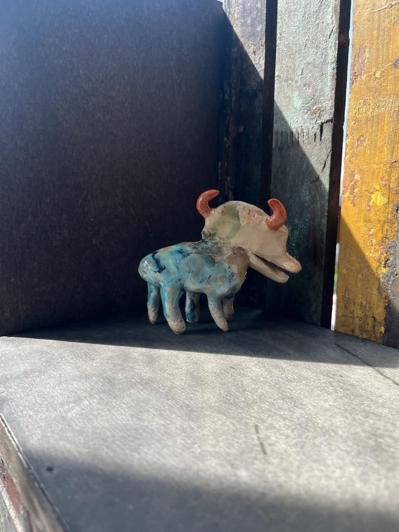 K&ouml;nnte beinhalten: Eine kleine Keramik-Tierfigur mit vier Beinen, einer langen Schnauze und zwei gebogenen orangefarbenen H&ouml;rnern. Der K&ouml;rper weist eine gesprenkelte Glasur in Blau-, Gr&uuml;n- und Cremet&ouml;nen auf, w&auml;hrend der Kopf haupts&auml;chlich cremefarben ist.