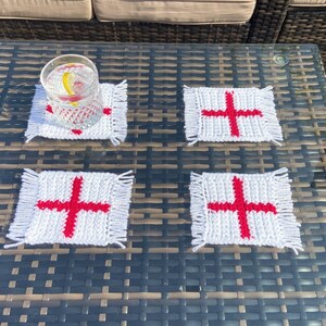 May include: Four white crocheted coasters with red crosses, similar to the English flag, on a brown wicker table. A glass with a lemon wedge and ice is on one of the coasters.