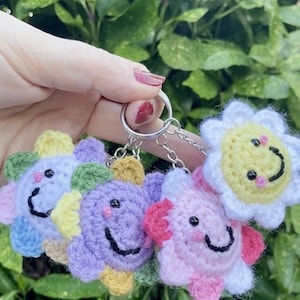 May include: A set of four crocheted flower keychains. Each keychain features a smiling face and is made in different colors, including yellow, white, purple, pink, green, and blue.