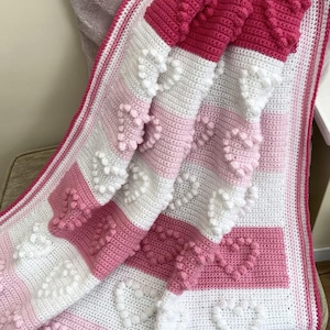 May include: A pink, white, and light pink crocheted blanket with a heart pattern. The blanket is folded and draped over a chair.