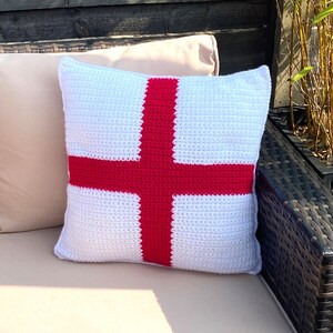 May include: A white crocheted cushion with a red cross design, resembling the flag of England.