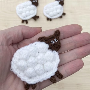 May include: Handmade crochet sheep with white wool and brown faces and legs. The sheep are small, with a textured, puffy appearance. Two additional sheep are visible in the background. The sheep are likely intended for crafting or decoration.