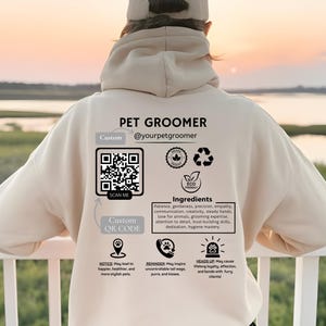 May include: A white hoodie with the text "PET GROOMER @yourpetgroomer" printed on the front. The hoodie also features a custom QR code, a logo with the text "ECO FRIENDLY", and a list of "Ingredients" for pet grooming services. The text includes: "Patience, gentleness, precision, empathy, communication, creativity, steady hands, love for animals, grooming expertise, attention to detail, trust-building skills, dedication, hygiene mastery."