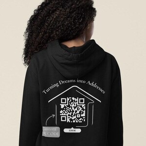Könnte beinhalten: Schwarzer Hoodie mit einem weißen Aufdruck, der "Turning Dreams into Addresses" lautet und ein Haus mit einem QR-Code darin zeigt. Der Text "Custom QR Code" und "Scan Me" ist ebenfalls im Aufdruck enthalten.