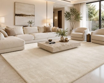 Puede incluir: Una alfombra grande, rectangular y de color crema sólido con una textura suave y afelpada que cubre el suelo de una sala de estar. La alfombra presenta un tono neutro uniforme que se extiende por toda la superficie visible del suelo.