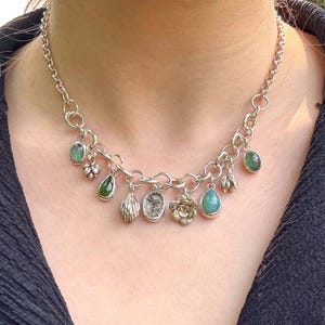 Könnte beinhalten: Silberfarbene Halskette mit verschiedenen Anhängern, darunter Edelsteine und florale Designs. Die Halskette hat eine Kette mit mehreren Anhänger-Anhängern in verschiedenen Formen und Farben, darunter Grün, Silber und Gold. Die Halskette wird um den Hals getragen.