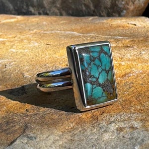 May include: A silver ring featuring a rectangular turquoise stone set in a bezel setting. The turquoise is a vibrant blue-green colour with brown matrix.