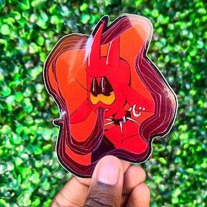 Puede incluir: Una pegatina de dibujos animados roja con un personaje parecido a un diablo con una expresi&oacute;n triste. El personaje tiene cuernos, un collar de p&uacute;as y un peque&ntilde;o pendiente. La pegatina tiene un borde blanco y se sostiene sobre un fondo verde y frondoso.