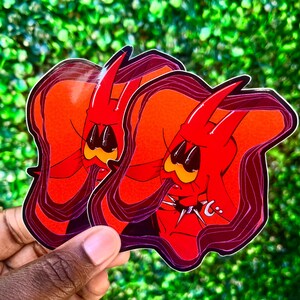Puede incluir: Dos pegatinas con un diablo de dibujos animados con piel roja, cuernos y boca amarilla. El diablo tiene una expresi&oacute;n triste y est&aacute; rodeado por un contorno rojo oscuro. Las pegatinas se sostienen sobre un fondo verde.