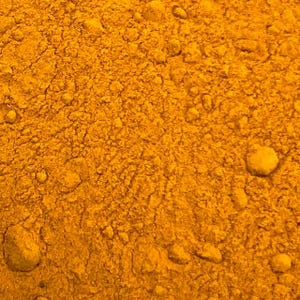 May include: Close-up of a textured, golden-yellow powder with a granular appearance. The surface is uneven, with small, irregular clumps scattered throughout. The color is consistent, suggesting a single substance or a finely ground mixture. The image highlights the texture and color of the powder.