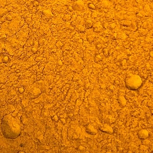 May include: Close-up of a textured, golden-yellow powder with a granular appearance. The surface is uneven, with small, irregular clumps scattered throughout. The color is consistent, suggesting a single substance or a finely ground mixture. The image highlights the texture and color of the powder.