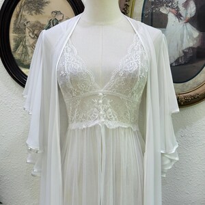 May include: A white sheer robe and matching chemise set. The robe features wide, layered flutter sleeves. The chemise has a V-neckline with floral lace detailing, a fitted lace bodice with three small buttons, and a flowing, sheer skirt.