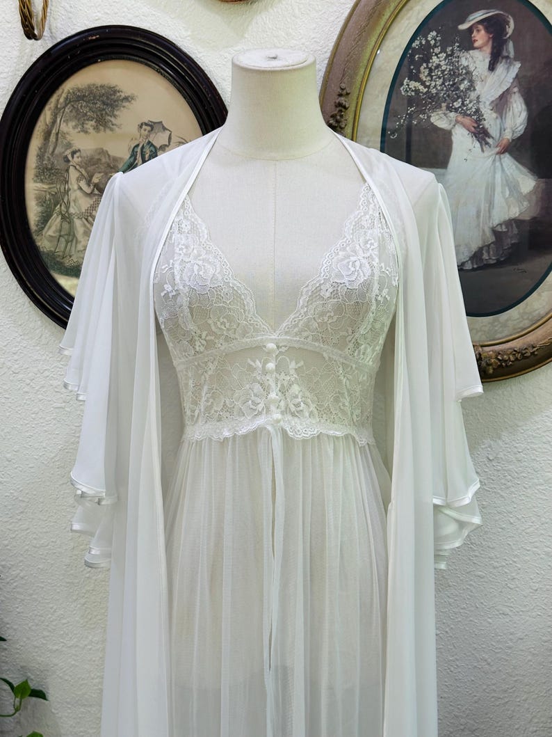 May include: A white sheer robe and matching chemise set. The robe features wide, layered flutter sleeves. The chemise has a V-neckline with floral lace detailing, a fitted lace bodice with three small buttons, and a flowing, sheer skirt.