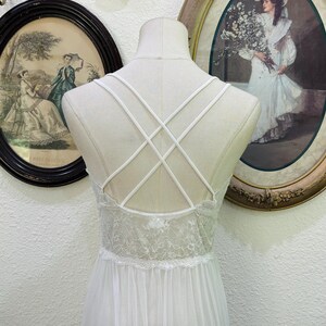 May include: A white nightgown featuring a sheer lace bodice with a scalloped edge and a flowing skirt. The back design includes thin, crisscrossing straps that form an X pattern, providing a delicate look against the solid white fabric.