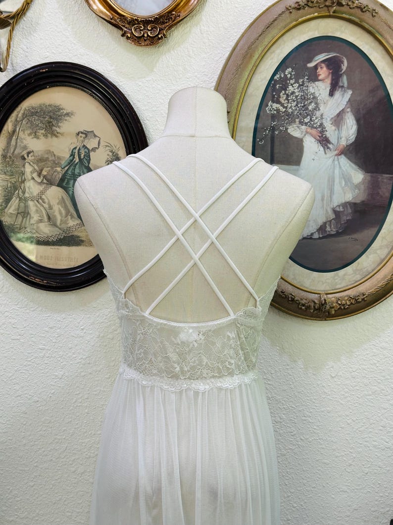 May include: A white nightgown featuring a sheer lace bodice with a scalloped edge and a flowing skirt. The back design includes thin, crisscrossing straps that form an X pattern, providing a delicate look against the solid white fabric.