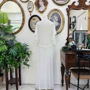 May include: A floor-length white sheer robe featuring short flutter sleeves, a thin waist tie, and a crisscross strap design on the back. The garment is displayed on a mannequin, showcasing a lightweight, flowing silhouette with a clean, solid white finish.