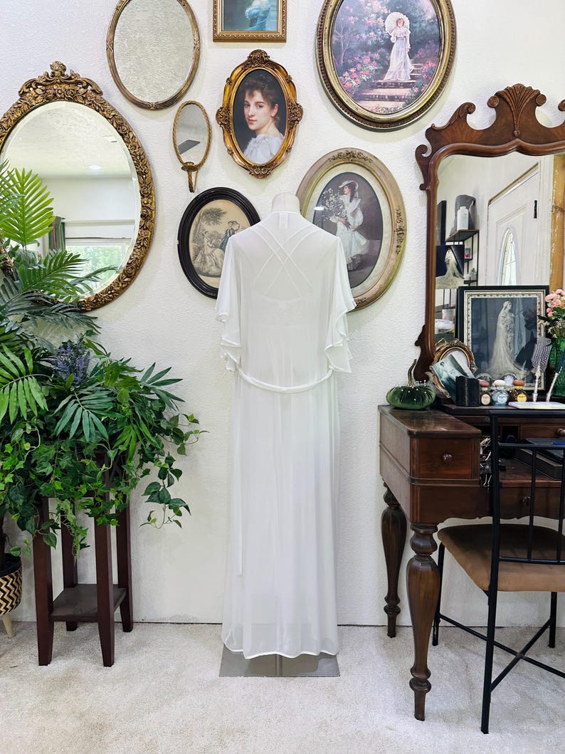 May include: A floor-length white sheer robe featuring short flutter sleeves, a thin waist tie, and a crisscross strap design on the back. The garment is displayed on a mannequin, showcasing a lightweight, flowing silhouette with a clean, solid white finish.