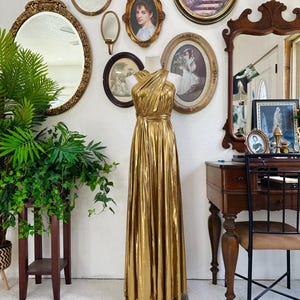 May include: A full-length, gold-colored evening gown on a mannequin. The dress features a draped, asymmetrical neckline and a flowing skirt. The background includes antique mirrors and framed portraits, suggesting a vintage or retro theme.