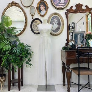 May include: A floor-length white sleeveless gown featuring a sheer overlay, a fitted bodice with delicate lace detailing, and a high-waisted design that flows into a long skirt with a central front slit, displayed on a mannequin stand.
