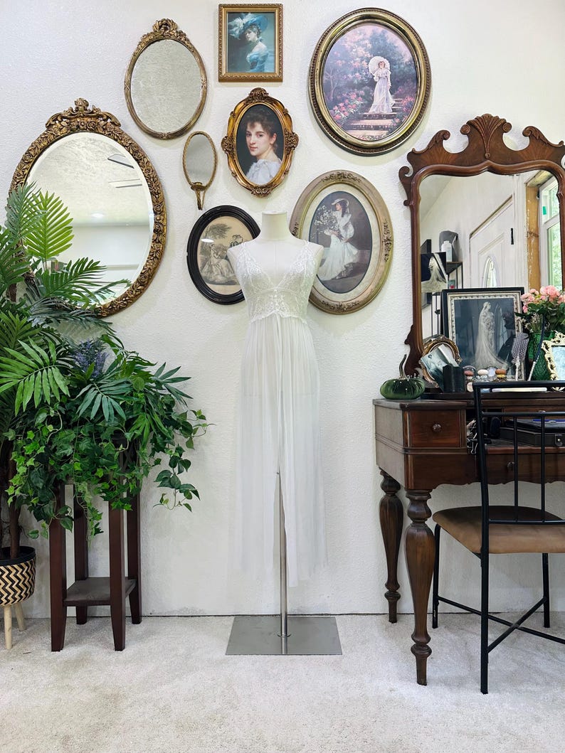 May include: A floor-length white sleeveless gown featuring a sheer overlay, a fitted bodice with delicate lace detailing, and a high-waisted design that flows into a long skirt with a central front slit, displayed on a mannequin stand.