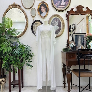 May include: A white floor-length robe with sheer, flowing sleeves and a matching sleeveless nightgown featuring a lace bodice. The garment is displayed on a mannequin, showcasing a lightweight, layered design with a clean, solid white color palette.