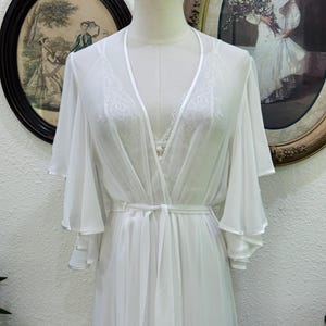 May include: A white sheer robe featuring wide, layered bell sleeves and a matching tie belt at the waist. The robe has a deep V-neckline with satin trim and is layered over a white camisole with delicate lace detailing along the neckline.