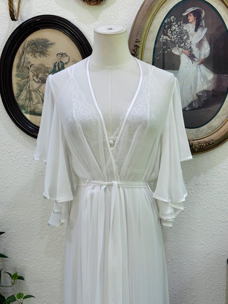 May include: A white sheer robe featuring wide, layered bell sleeves and a matching tie belt at the waist. The robe has a deep V-neckline with satin trim and is layered over a white camisole with delicate lace detailing along the neckline.