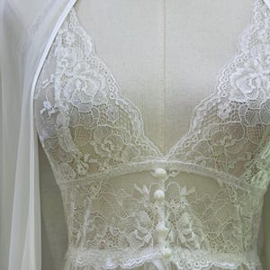 May include: A white lace camisole featuring a deep V-neckline, floral lace detailing, and a front button placket with four small round buttons. The garment is layered under a matching sheer white robe with thin straps, displayed on a mannequin.