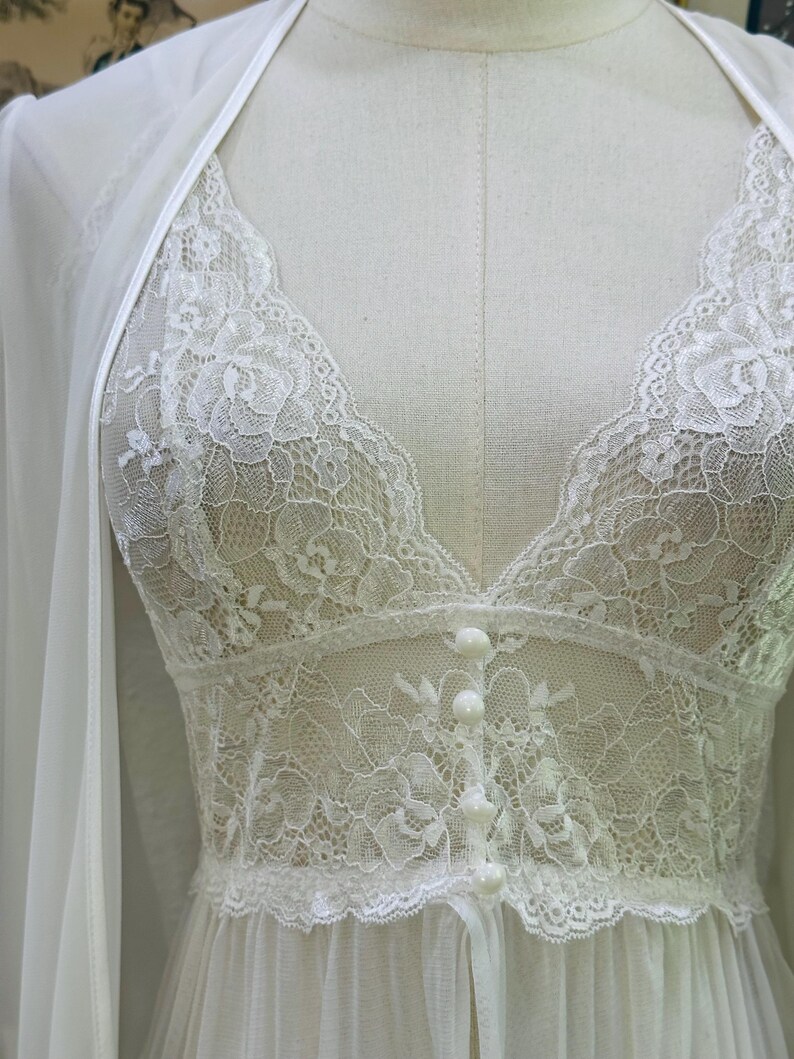 May include: A white lace camisole featuring a deep V-neckline, floral lace detailing, and a front button placket with four small round buttons. The garment is layered under a matching sheer white robe with thin straps, displayed on a mannequin.