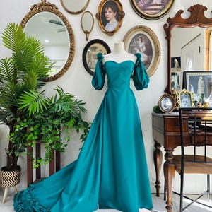 May include: A teal-colored formal gown with a sweetheart neckline and puffed sleeves. The dress features a flowing skirt with a ruffled hem. The dress is displayed on a mannequin in front of a wall decorated with antique mirrors and framed artwork.