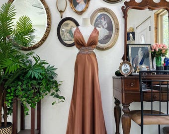 May include: A floor-length, copper-colored satin gown on a mannequin, featuring a plunging neckline and a beaded belt. The dress is displayed in a room with antique mirrors and framed vintage portraits, creating a vintage aesthetic.