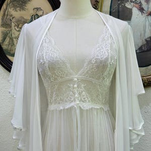May include: A white sheer robe and matching nightgown set. The robe features wide, layered flutter sleeves. The nightgown has a V-neckline, floral lace bodice detailing, and a row of small white buttons down the center front, flowing into a sheer skirt.