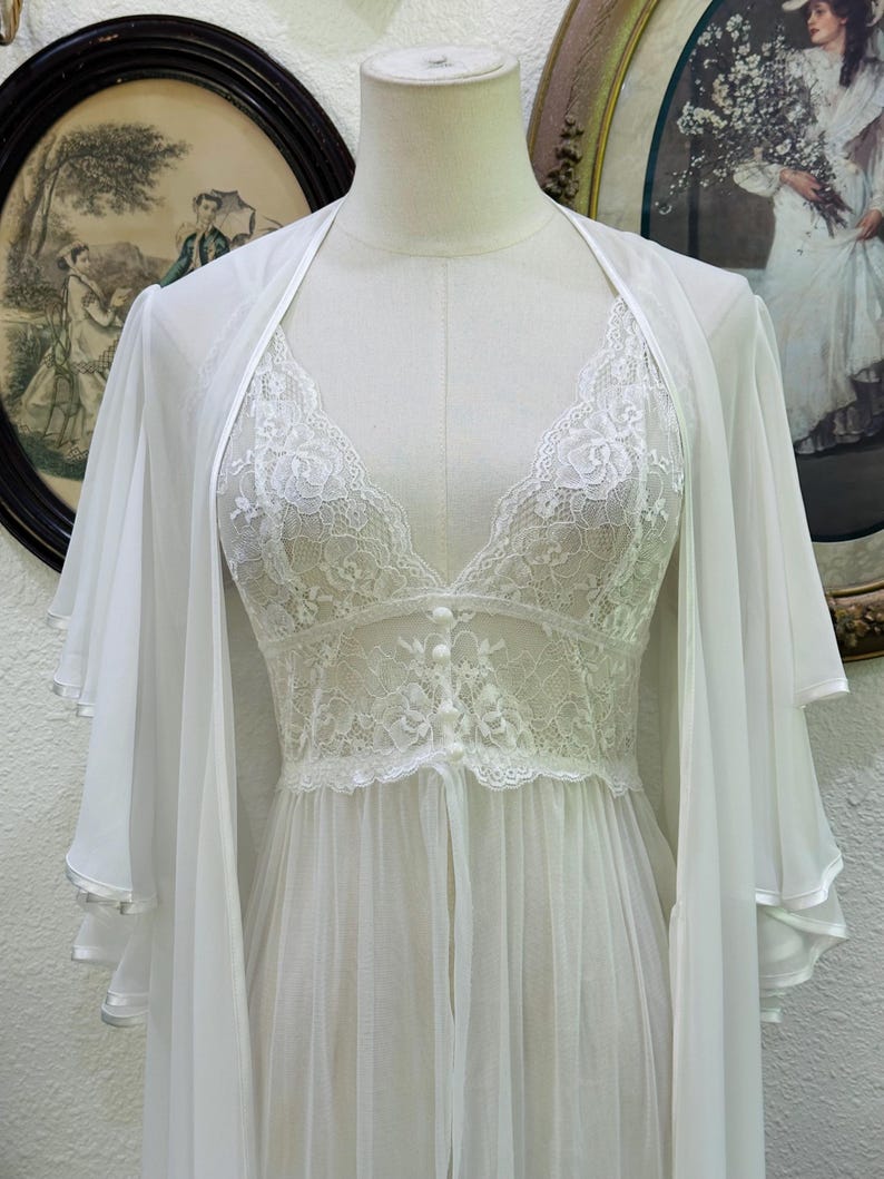 May include: A white sheer robe and matching nightgown set. The robe features wide, layered flutter sleeves. The nightgown has a V-neckline, floral lace bodice detailing, and a row of small white buttons down the center front, flowing into a sheer skirt.
